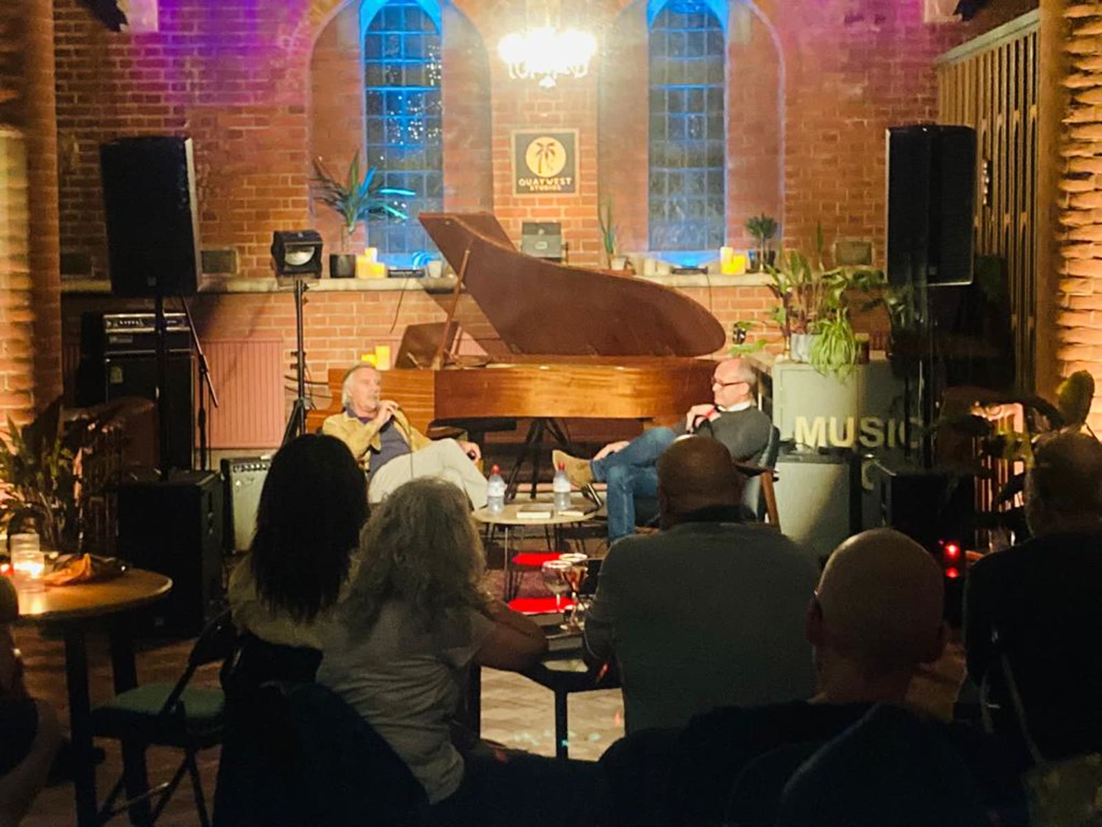 Peter Stoneman interviewing Glen Matlock on stage beside a grand piano, with an audience in the foreground.